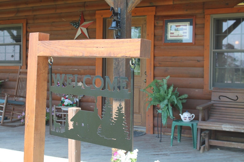 May include: A wooden sign with a metal cutout that says "WELCOME" with a cabin and trees in the background. The sign is hanging from a wooden post in front of a log cabin.
