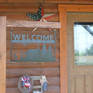 May include: A metal welcome sign with a cabin and trees silhouette hangs from a wooden post in front of a log cabin. The sign is black and the cabin is brown. The sign says "WELCOME" in large letters. A red, white, and blue star is hanging above the sign.