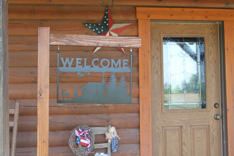 May include: A metal welcome sign with a cabin and trees silhouette hangs from a wooden post in front of a log cabin. The sign is black and the cabin is brown. The sign says "WELCOME" in large letters. A red, white, and blue star is hanging above the sign.