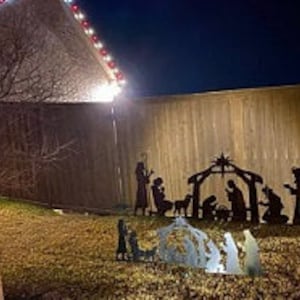 May include: Metal silhouette of the nativity scene with the figures of Mary, Joseph, baby Jesus, and the three wise men. The figures are black and stand against a wooden fence background.