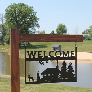 May include: A black metal welcome sign with a cabin, trees, and a deer silhouette. The sign is hanging from a wooden post in front of a lake and a sandy beach.
