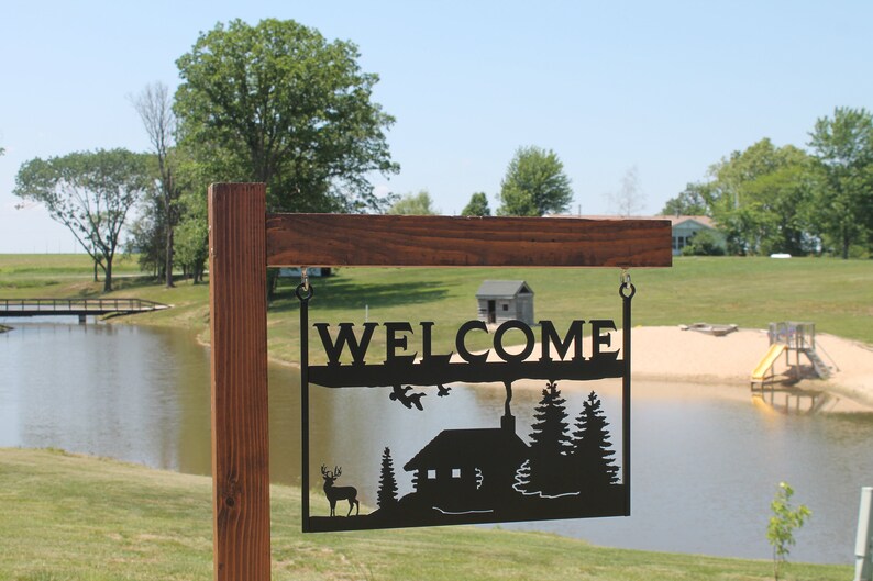 May include: A black metal welcome sign with a cabin, trees, and a deer silhouette. The sign is hanging from a wooden post in front of a lake and a sandy beach.
