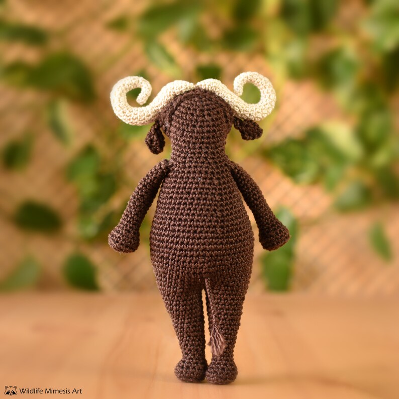 K&ouml;nnte beinhalten: Ein handgefertigtes, braunes geh&auml;keltes B&uuml;ffeltier mit cremefarbenen H&ouml;rnern. Das Amigurumi-Tier hat einen kleinen Schwanz und steht auf einer Holzoberfl&auml;che. Der Hintergrund ist mit gr&uuml;nen Bl&auml;ttern verschwommen.