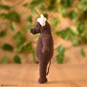 K&ouml;nnte beinhalten: Geh&auml;kelte braune Tierfigur mit wei&szlig;em Hut. Das Tier hat eine schwarze Nase und einen kleinen Schwanz. Die Figur steht auf einer Holzoberfl&auml;che, im Hintergrund gr&uuml;nes Laub. Ein einzigartiges handgefertigtes Objekt.