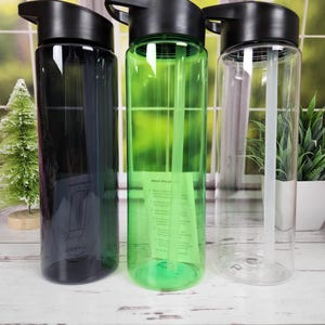 May include: Three clear plastic water bottles with black lids. One bottle is black, one is green, and one is clear. Each bottle has a straw inside. The bottles are on a white wooden surface.