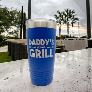 May include: A blue stainless steel tumbler with a silver lid. The tumbler has the text "Daddy's gonna grill" engraved on the side.