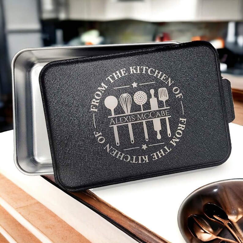 May include: A black baking pan with a removable bottom and a personalized inscription that reads "From the Kitchen of Alexis McCabe From the Kitchen of". The inscription is surrounded by a circle with a star on each side and a graphic of kitchen utensils.