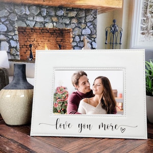 May include: White picture frame with a stitched border and the text "love you more" in cursive with a small heart. The frame has a photo of a couple in front of a Christmas tree.