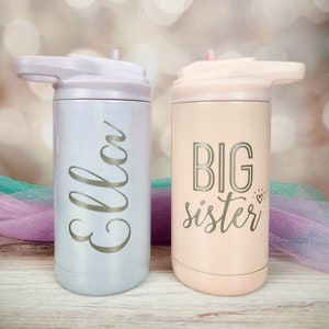 May include: Two stainless steel water bottles with lids and handles. The bottle on the left is light blue with the name "Ella" written on it. The bottle on the right is pink with the text "BIG sister" written on it.