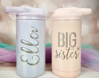 May include: Two stainless steel tumblers with lids and straws. The left tumbler is light purple with a glittery finish and the name Ella in silver script. The right tumbler is light peach with a glittery finish and the text BIG sister in silver.