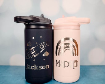 May include: A dark navy blue insulated water bottle with a rocket ship and stars engraving and the name Jackson, next to a light pink insulated water bottle with a rainbow engraving and the name Madelyn, both featuring black and pink flip-top lids.