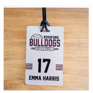 May include: A rectangular tag with the text "ROSSFORD BULLDOGS VOLLEYBALL" in maroon and white. Below is the number "17" and the name "EMMA HARRIS" in black. A black cord is attached at the top.