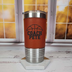 May include: A stainless steel travel mug with a brown basketball-textured sleeve. The sleeve has a basketball graphic and the text "COACH PETE" in black.