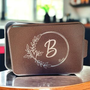 May include: A brown metal baking pan with a white engraved design of a wreath with a heart and the letter B inside. The text "Brown" is also engraved on the pan.
