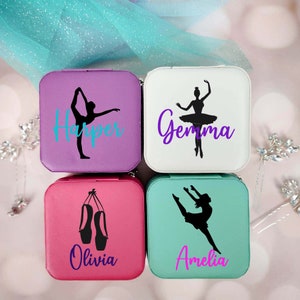 May include: Four square jewelry boxes with different designs. The boxes are purple, white, pink, and teal. The designs include a ballerina silhouette, a pair of ballet shoes, and the names "Harper", "Gemma", "Olivia", and "Amelia".