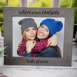 May include: A gray picture frame with a stitched border and the text "The love between cousins lasts forever". The frame features a photo of two young women wearing hats, smiling and hugging.