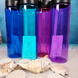 May include: A collection of colorful water bottles in blue, teal, purple, and magenta, each with a black lid. The bottles are displayed with seashells on a sandy surface. The bottles have a capacity of 24 oz.