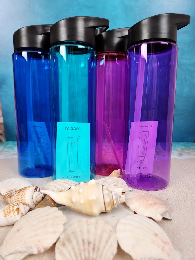 May include: A collection of colorful water bottles in blue, teal, purple, and magenta, each with a black lid. The bottles are displayed with seashells on a sandy surface. The bottles have a capacity of 24 oz.