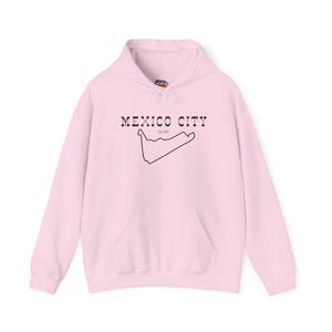 May include: A light pink hoodie with a front pocket and a drawstring hood. The front of the hoodie features the text "MEXICO CITY" above a black outline of a race track.