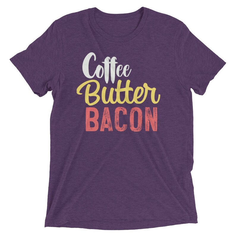 May include: Purple short-sleeved t-shirt with the text "Coffee Butter Bacon" in a vintage style font. The words "Coffee" and "Butter" are in yellow, and the word "Bacon" is in pink.