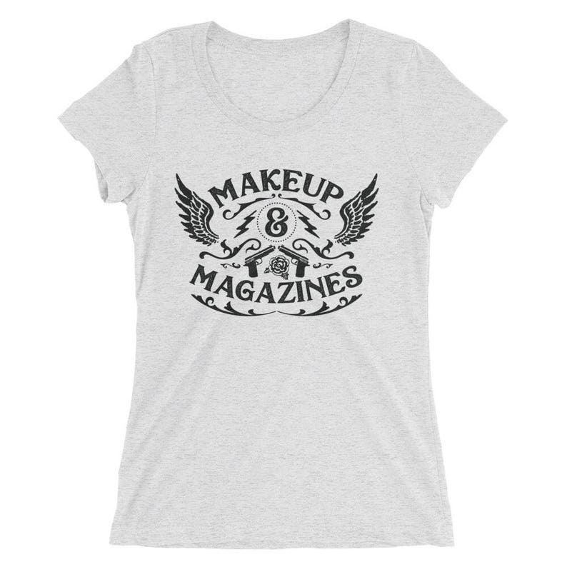Puede incluir: Una camiseta gris jaspeada para mujer con un dise&ntilde;o gr&aacute;fico negro. El gr&aacute;fico presenta un par de pistolas cruzadas con una rosa y el texto "MAKEUP & MAGAZINES".