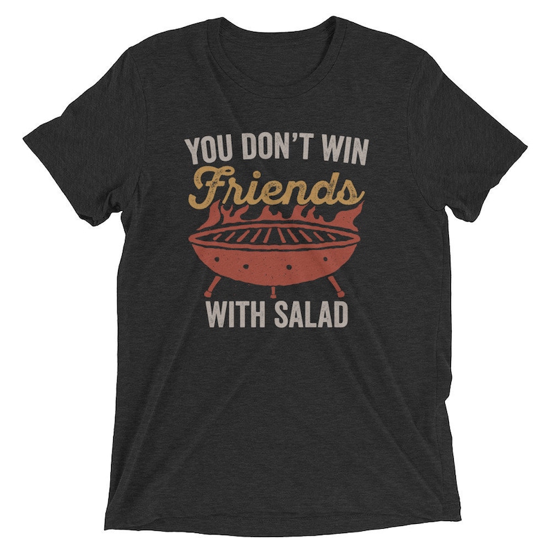 May include: A dark grey t-shirt with a vintage-style graphic of a grill and the text "You don't win friends with salad."