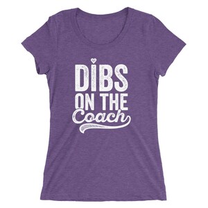 Puede incluir: Camiseta morada con la frase blanca "Dibs on the Coach", con un peque&ntilde;o coraz&oacute;n sobre la "i" de "Dibs". La camiseta tiene cuello redondo y mangas cortas. La tela tiene una textura jaspeada.
