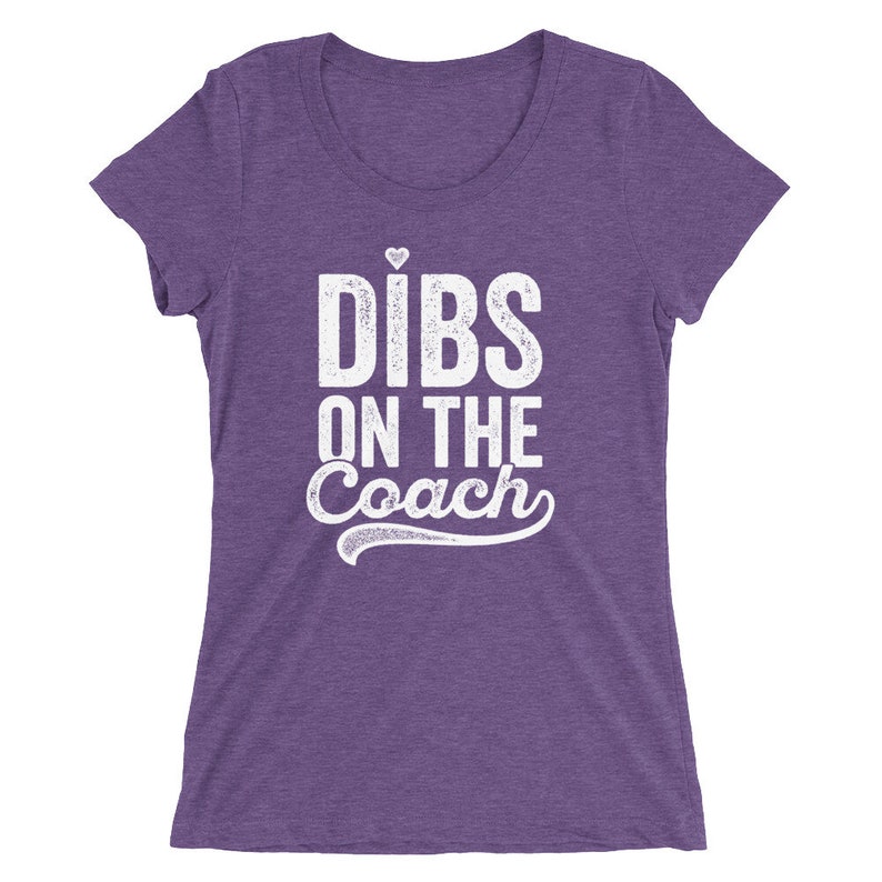 Puede incluir: Camiseta morada con la frase blanca "Dibs on the Coach", con un peque&ntilde;o coraz&oacute;n sobre la "i" de "Dibs". La camiseta tiene cuello redondo y mangas cortas. La tela tiene una textura jaspeada.