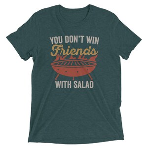 May include: A teal blue t-shirt with the text "You don't win friends with salad" in white and yellow. A red and orange grill with flames is in the center of the text.