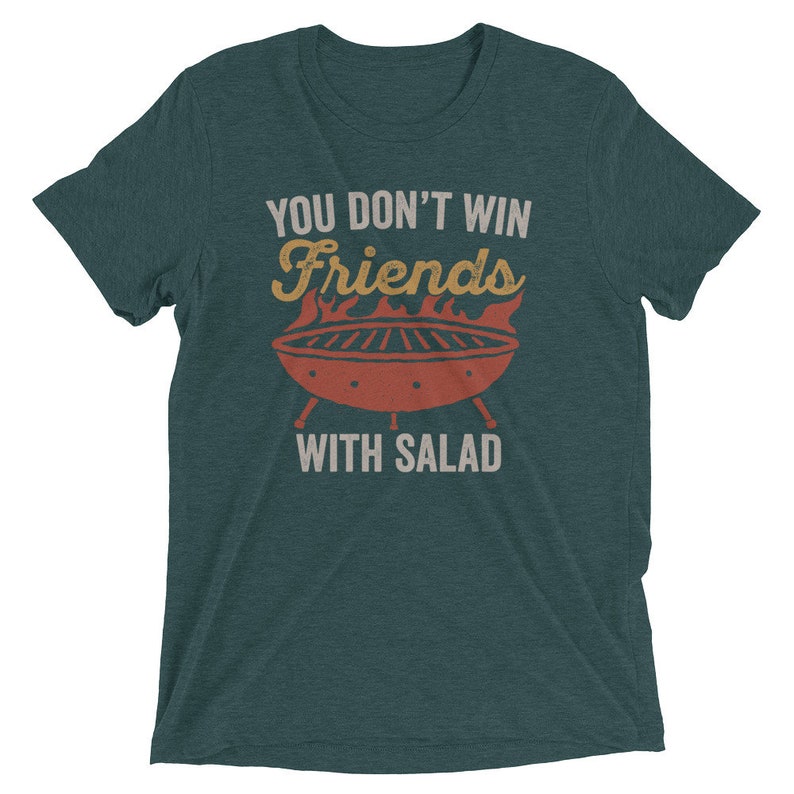 May include: A teal blue t-shirt with the text "You don't win friends with salad" in white and yellow. A red and orange grill with flames is in the center of the text.