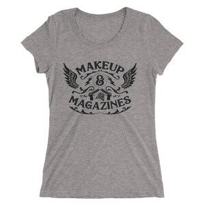 Puede incluir: Camiseta gris con un dise&ntilde;o gr&aacute;fico negro que dice "MAKEUP & MAGAZINES" con un dise&ntilde;o estilizado de alas, pistolas y una rosa.