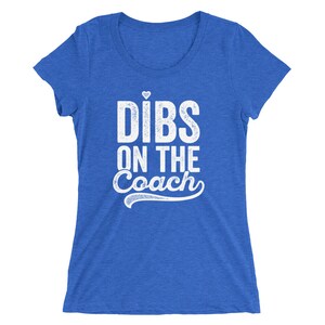 Puede incluir: Una camiseta azul con la frase blanca "DIBS ON THE COACH", con un peque&ntilde;o coraz&oacute;n encima de la palabra "DIBS". El texto tiene un aspecto desgastado. La camiseta tiene mangas cortas y cuello redondo, t&iacute;pico de una prenda informal.