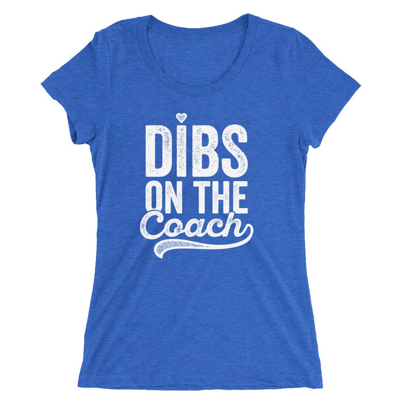 Puede incluir: Una camiseta azul con la frase blanca "DIBS ON THE COACH", con un peque&ntilde;o coraz&oacute;n encima de la palabra "DIBS". El texto tiene un aspecto desgastado. La camiseta tiene mangas cortas y cuello redondo, t&iacute;pico de una prenda informal.