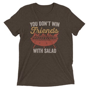 May include: A brown t-shirt with the text "You don't win friends with salad" in white. The text is above a red and orange illustration of a grill with flames.