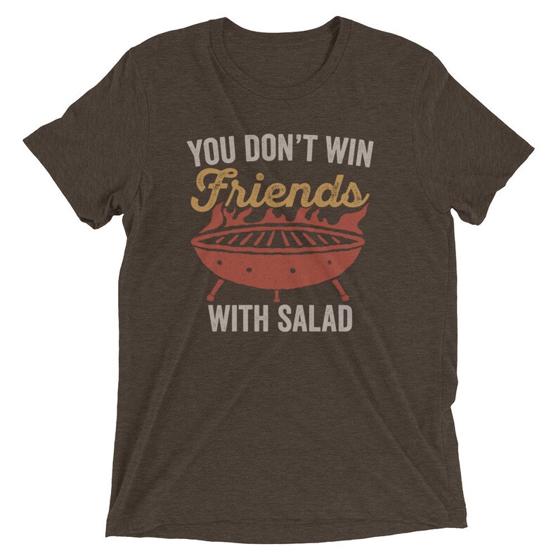May include: A brown t-shirt with the text "You don't win friends with salad" in white. The text is above a red and orange illustration of a grill with flames.