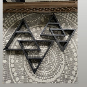 May include: Size comparison between the original double triangle and the XL double triangle. Two black wooden geometric triangle shelves are mounted on a wall. The larger shelf is twice the size of the smaller shelf.