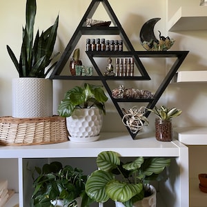 May include: A black triangular shelf unit with multiple shelves, displaying various items, including plants, crystals, and essential oils. The shelf is mounted on a white wall, and there are additional white shelves above and below it.