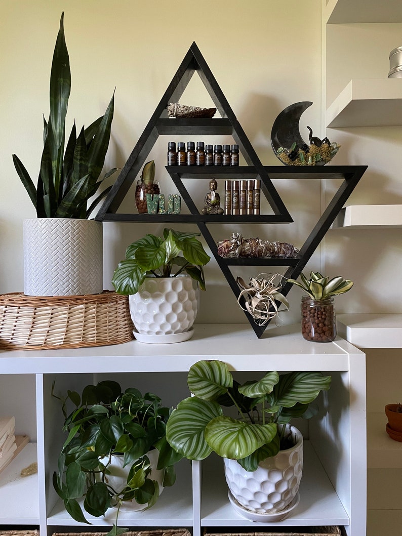 May include: A black triangular shelf unit with multiple shelves, displaying various items, including plants, crystals, and essential oils. The shelf is mounted on a white wall, and there are additional white shelves above and below it.