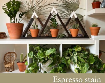 May include: A wooden shelf unit with a mountain range design, stained in espresso. The shelf is filled with potted plants, including succulents and a small potted tree. The shelf is mounted on a white wall.