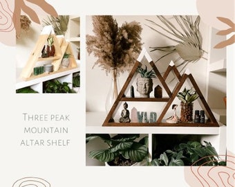 May include: A wooden shelf shaped like three mountain peaks with white painted peaks. The shelf has three levels and is decorated with plants, crystals, and figurines. The shelf is labeled "Three Peak Mountain Altar Shelf".