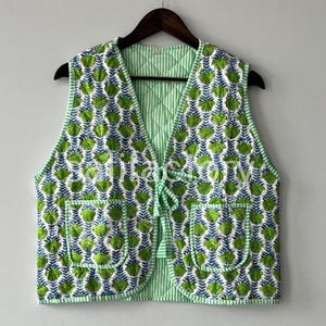 May include: A quilted vest featuring a green and white floral design and two front pockets. The vest has a striped green lining and a tie closure. It is suspended from a wooden hanger.