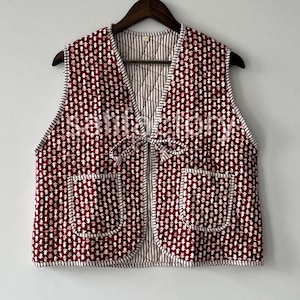 Puede incluir: Un chaleco acolchado sin mangas con un estampado floral burdeos y blanco. El chaleco tiene dos bolsillos delanteros y un cierre de lazo. El forro interior es un estampado de rayas. El chaleco está colgado de una percha de madera.