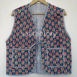 Könnte beinhalten: Eine ärmellose Weste mit einem Blumenmuster in Rosa und Rot auf blauem Grund. Die Weste hat zwei Taschen und ein gestreiftes Futter. Sie wird vorne mit einer passenden Stoffkrawatte gebunden. Die Weste hängt an einem Holzbügel.