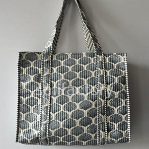 Puede incluir: Un bolso tote acolchado con un patrón repetitivo en forma de abanico en tonos grises y crema. El bolso tiene asas y ribetes a juego con un diseño de rayas. El bolso es rectangular y parece estar hecho de tela.