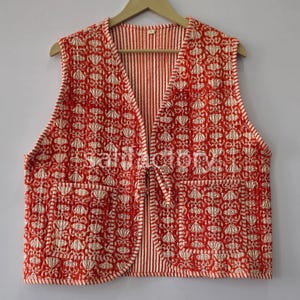 May include: A red and white quilted vest with a floral pattern. The vest has a V-neck, two front pockets, and a tie closure. The interior lining is striped. The vest is hanging on a wooden hanger.
