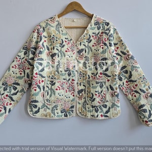 May include: Cream quilted jacket with a floral design in red, green, and blue. The jacket features a V-neck, two pockets, and long sleeves. It is displayed on a wooden hanger.