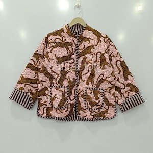 May include: A quilted jacket featuring a repeating pattern of brown tigers on a pink background with small red dots. The jacket has a black and white striped collar, cuffs, and pocket trim. The jacket is hanging on a white wall.