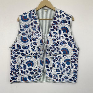 May include: A sleeveless vest with a blue and white floral pattern and a striped interior. The vest has a tie closure and two pockets. The design features blue and red accents on a white background. The vest is hanging on a wooden hanger.