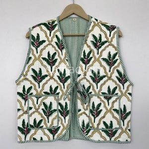 May include: A quilted vest with a white background and a repeating pattern of green leaves and red accents. The vest has two pockets and a tie closure. The interior lining is striped green and white.