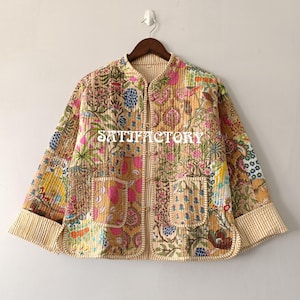 May include: A patchwork quilted jacket with floral and botanical patterns in pink, blue, yellow, and green. The jacket has a mandarin collar, two front pockets, and a striped trim. The word "SATISFACTORY" is printed on the front. The jacket is hung on a wooden hanger.
